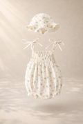 Floating baby romper set with matching sun hat in soft ivory fabric, featuring a delicate floral print, shown against a warm beige background with gentle natural light and soft shadows.